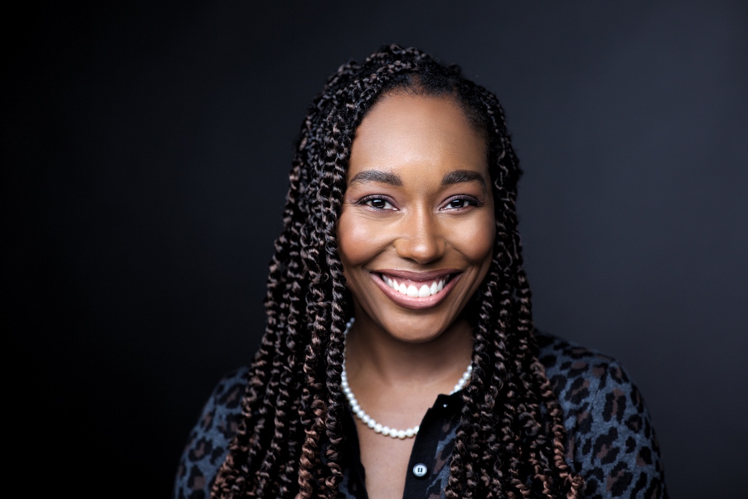 Corporate headshot NYC — smiling woman with braids in dark blouse, dark studio backdrop